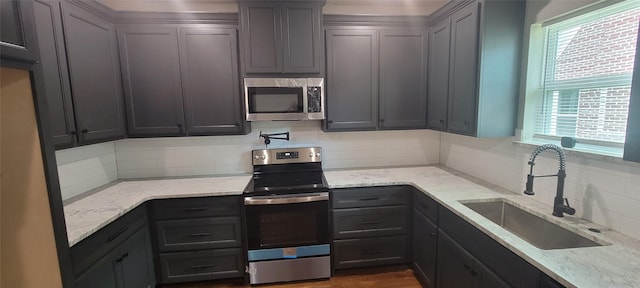 kitchen with appliances with stainless steel finishes, a sink, backsplash, light stone counters, and gray cabinets