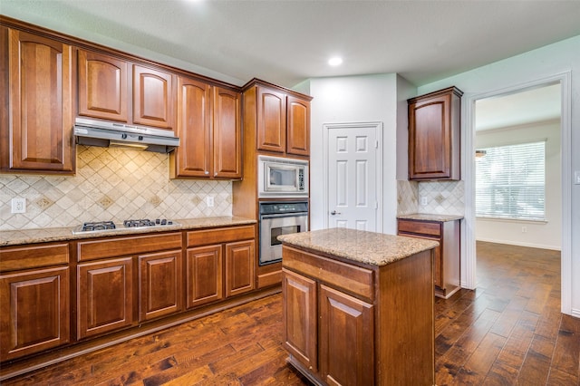 kitchen with appliances with stainless steel finishes, under cabinet range hood, dark wood-style flooring, light stone counters, and tasteful backsplash
