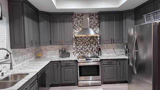 kitchen featuring appliances with stainless steel finishes, wall chimney range hood, a sink, a raised ceiling, and gray cabinetry