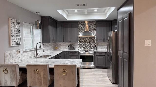 kitchen with wall chimney range hood, appliances with stainless steel finishes, a sink, a peninsula, and light countertops
