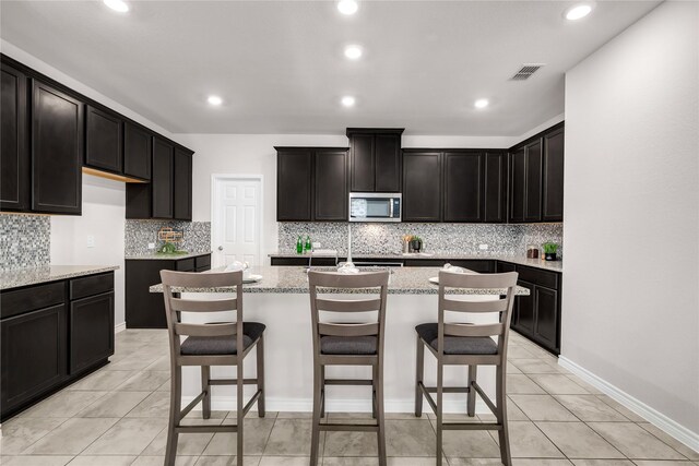 3433 Alamosa River Dr, Celina, TX 75078 - kitchen with stainless steel microwave, an island with sink, backsplash, light stone counters, and light tile patterned flooring