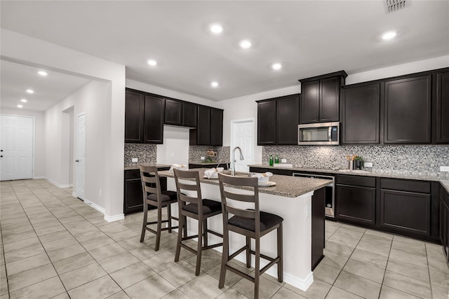 3433 Alamosa River Dr, Celina, TX 75078 - kitchen featuring stainless steel microwave, light tile patterned floors, decorative backsplash, recessed lighting, and a center island with sink