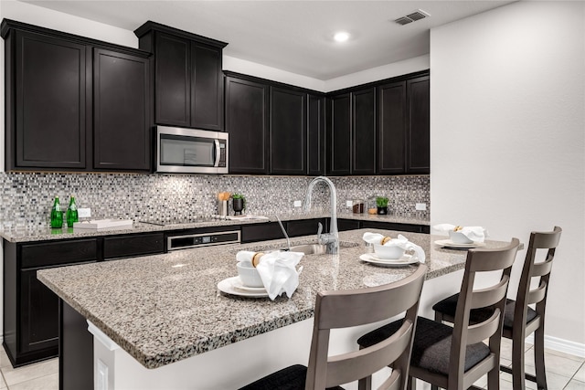 3433 Alamosa River Dr, Celina, TX 75078 - kitchen featuring stainless steel microwave, dark cabinetry, light tile patterned floors, and light stone counters