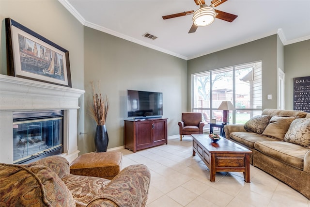 living room with ornamental molding, ceiling fan, light tile patterned floors, baseboards, and a glass covered fireplace