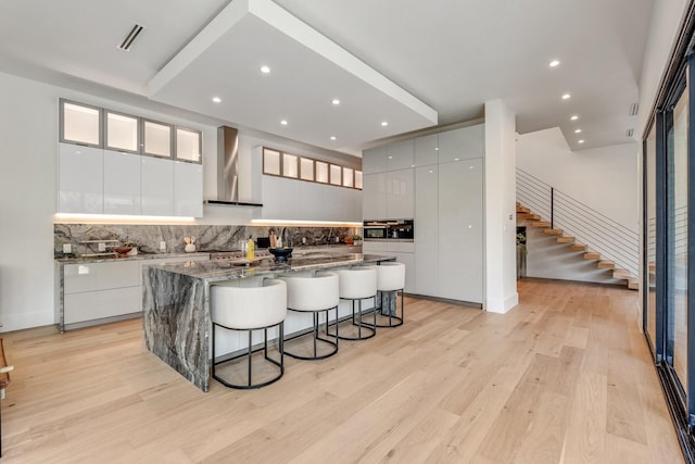 kitchen featuring wall chimney range hood, modern cabinets, a center island with sink, tasteful backsplash, and a kitchen breakfast bar