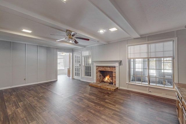 unfurnished living room featuring beam ceiling, a brick fireplace, a ceiling fan, dark wood-style flooring, and a decorative wall