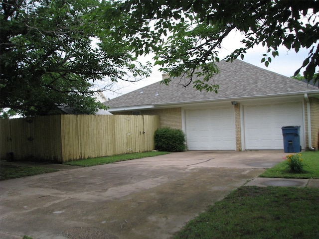 view of property exterior with concrete driveway, roof with shingles, fence, an attached garage, and brick siding