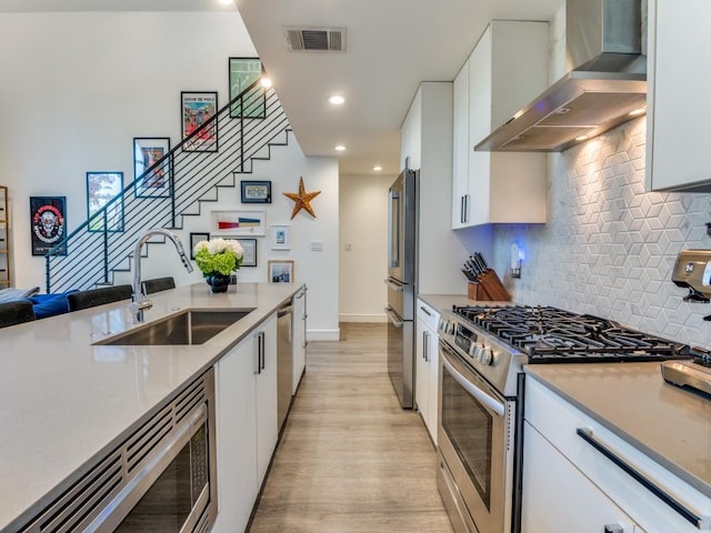 kitchen featuring wall chimney exhaust hood, stainless steel appliances, a sink, light wood-style flooring, and white cabinets