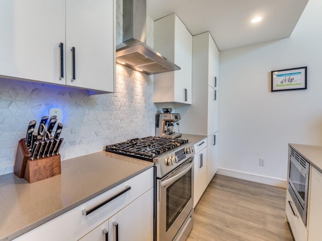 kitchen featuring wall chimney exhaust hood, appliances with stainless steel finishes, white cabinets, light wood finished floors, and decorative backsplash