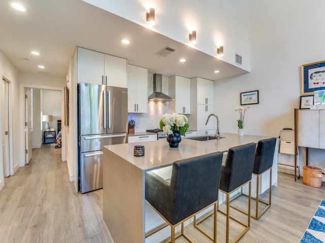 kitchen featuring freestanding refrigerator, wall chimney range hood, light wood-style floors, a center island with sink, and recessed lighting
