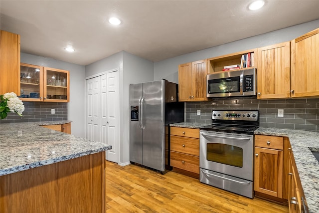 kitchen featuring stainless steel appliances, light wood-type flooring, tasteful backsplash, light stone counters, and glass insert cabinets