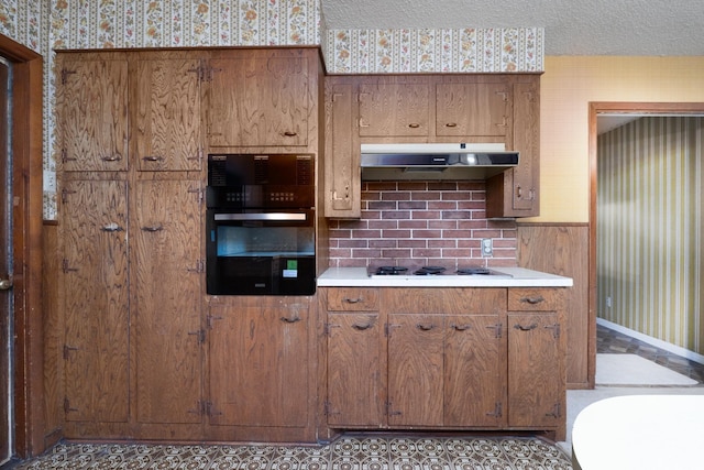 kitchen featuring under cabinet range hood, wallpapered walls, white electric cooktop, light countertops, and brown cabinets