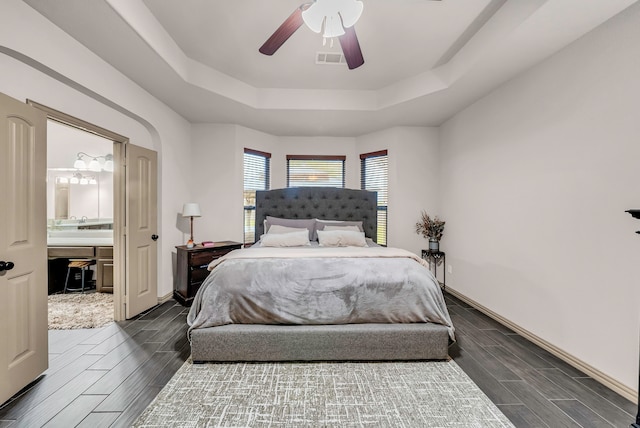 bedroom with a raised ceiling, wood finish floors, baseboards, ensuite bathroom, and a ceiling fan