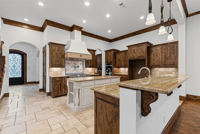 kitchen with custom exhaust hood, arched walkways, a center island with sink, ornamental molding, and backsplash
