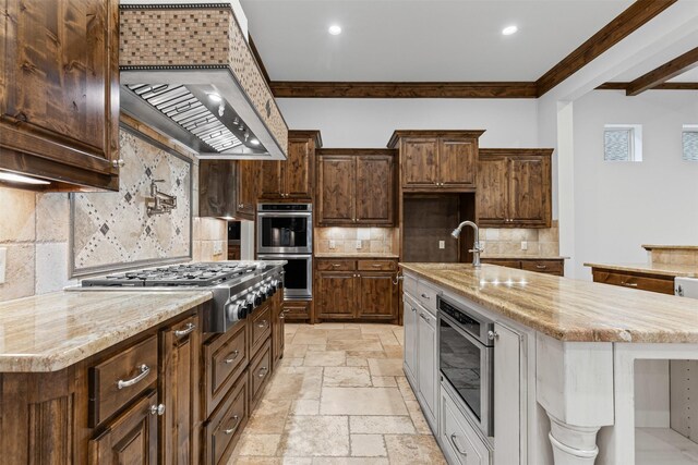kitchen with appliances with stainless steel finishes, ornamental molding, custom range hood, a kitchen island with sink, and stone tile flooring