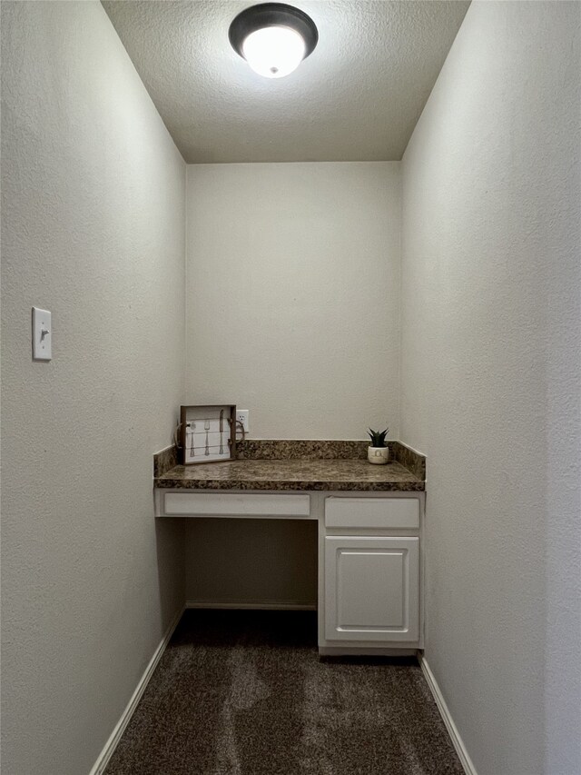 unfurnished office featuring built in desk, dark colored carpet, baseboards, and a textured ceiling