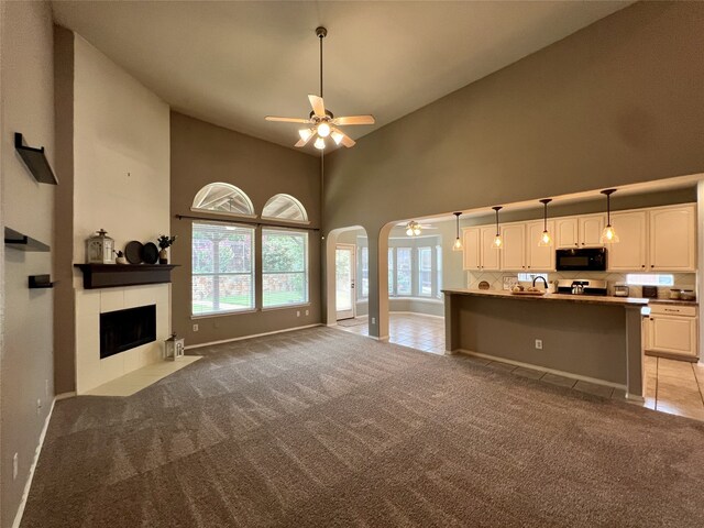 unfurnished living room with a ceiling fan, a towering ceiling, light carpet, arched walkways, and a tiled fireplace