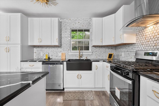 kitchen with appliances with stainless steel finishes, wall chimney exhaust hood, a sink, dark countertops, and white cabinets