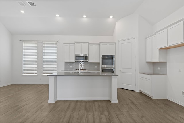 kitchen with appliances with stainless steel finishes, lofted ceiling, tasteful backsplash, light wood-type flooring, and a center island with sink