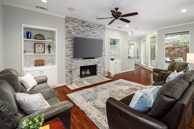 living room with ornamental molding, ceiling fan, built in shelves, dark wood-style flooring, and a stone fireplace