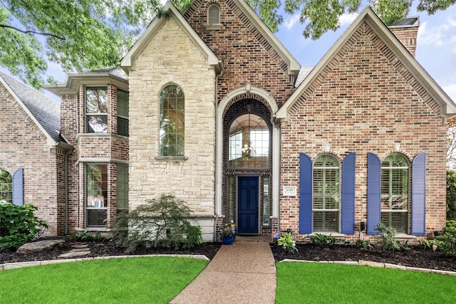 french country home featuring a front lawn, brick siding, a shingled roof, and stone siding