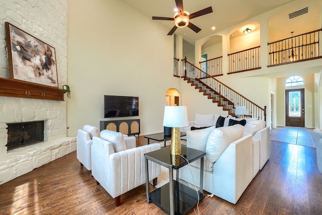 living room featuring a towering ceiling, dark wood-type flooring, stairs, ceiling fan, and a stone fireplace