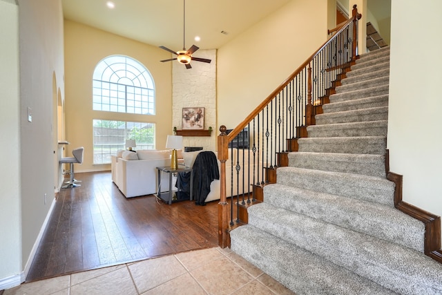 staircase featuring a high ceiling, a stone fireplace, ceiling fan, baseboards, and wood-type flooring
