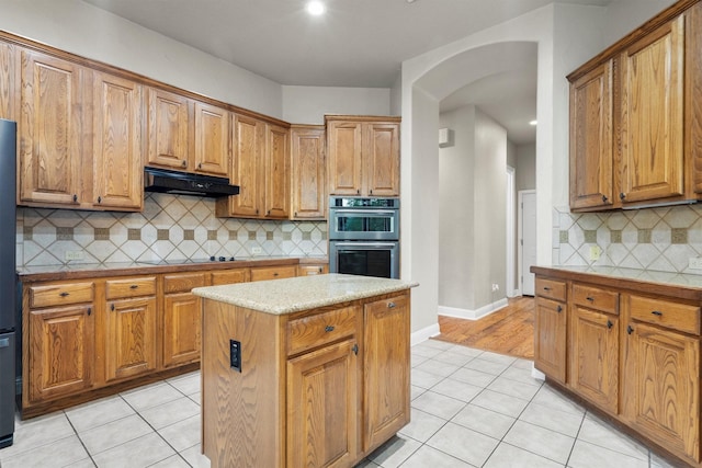 kitchen with under cabinet range hood, double oven, light tile patterned floors, arched walkways, and backsplash