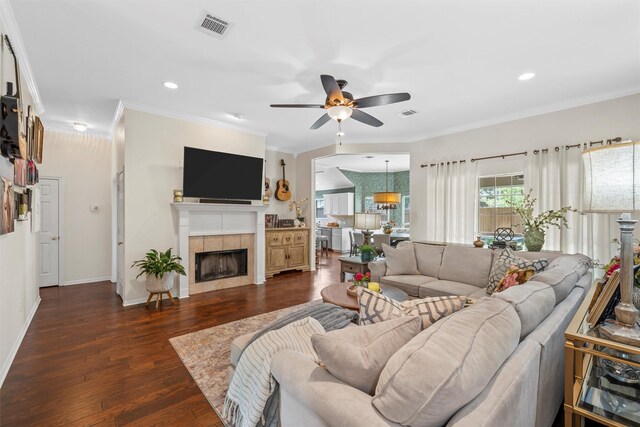 living room featuring ceiling fan, dark wood-style flooring, a tile fireplace, crown molding, and baseboards