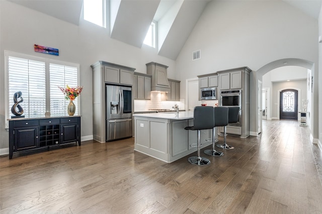 kitchen featuring stainless steel appliances, gray cabinets, high vaulted ceiling, arched walkways, and a breakfast bar area