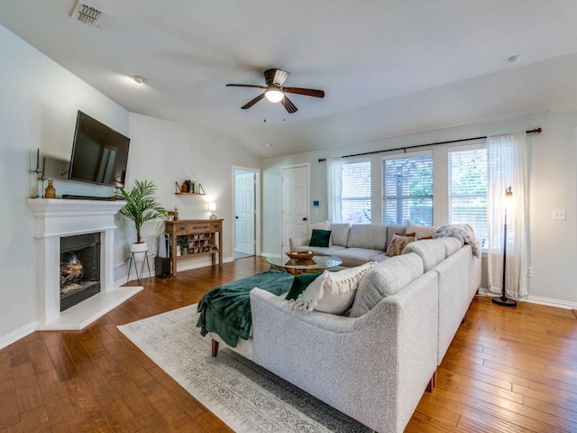 living room with a ceiling fan, wood-type flooring, baseboards, a fireplace with raised hearth, and vaulted ceiling