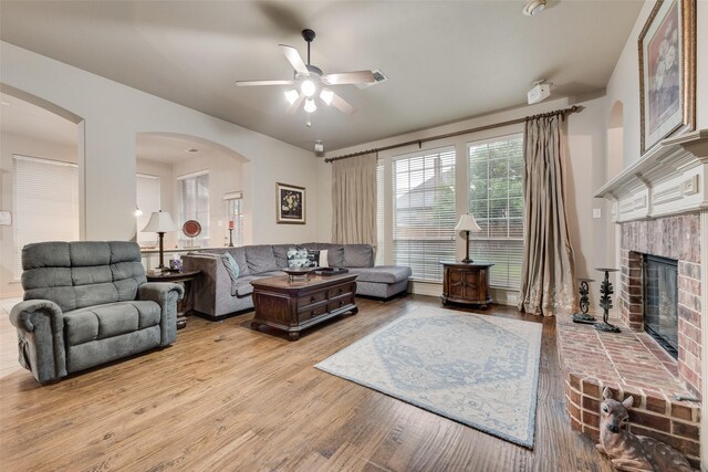 living area featuring a ceiling fan, light wood-type flooring, a brick fireplace, and arched walkways