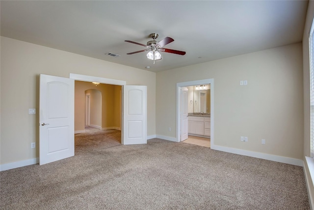 unfurnished bedroom featuring light colored carpet, arched walkways, baseboards, connected bathroom, and a ceiling fan