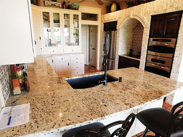 kitchen with appliances with stainless steel finishes, a sink, light stone counters, a breakfast bar area, and glass insert cabinets