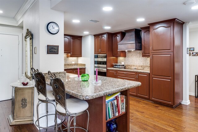 kitchen with premium range hood, stainless steel double oven, crown molding, wood finished floors, and backsplash