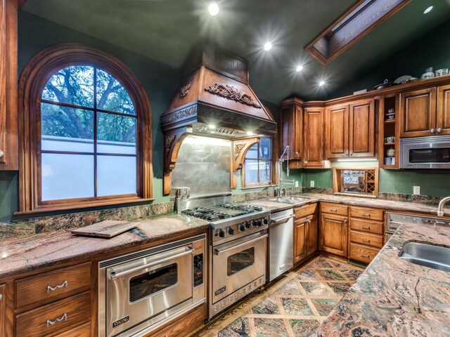 kitchen with appliances with stainless steel finishes, a sink, vaulted ceiling, brown cabinetry, and light stone counters