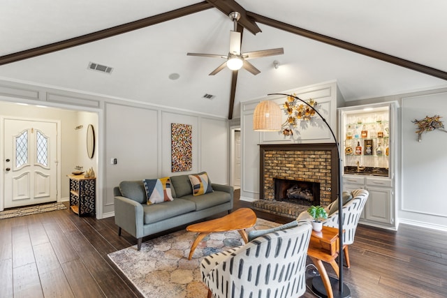 living room featuring a decorative wall, a ceiling fan, dark wood-style floors, a brick fireplace, and beamed ceiling