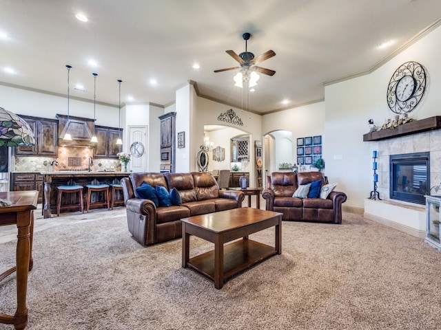 living area featuring light carpet, arched walkways, a ceiling fan, ornamental molding, and a tiled fireplace