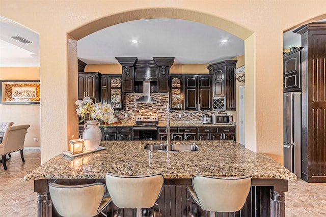 kitchen with arched walkways, stainless steel appliances, wall chimney range hood, light stone counters, and recessed lighting