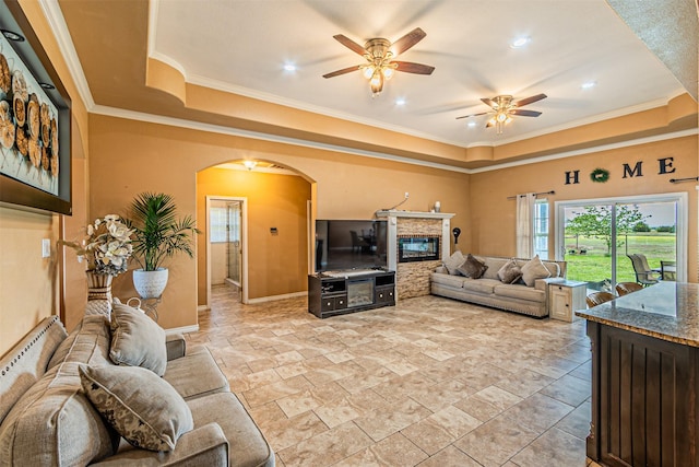 living room featuring a tray ceiling, ceiling fan, a fireplace, ornamental molding, and arched walkways