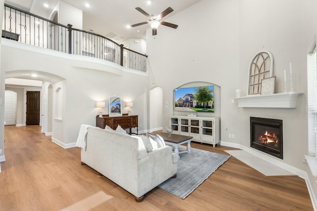living area featuring ceiling fan, a high ceiling, wood finished floors, a fireplace with flush hearth, and arched walkways