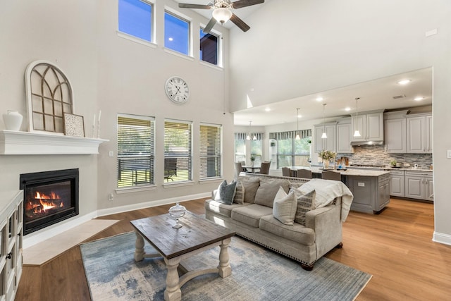 living room featuring a ceiling fan, a glass covered fireplace, light wood-style floors, a towering ceiling, and recessed lighting