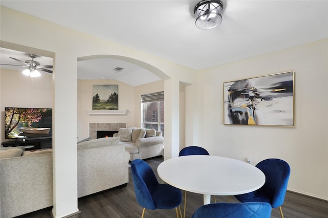 dining area with dark wood-type flooring, a tiled fireplace, arched walkways, a ceiling fan, and baseboards