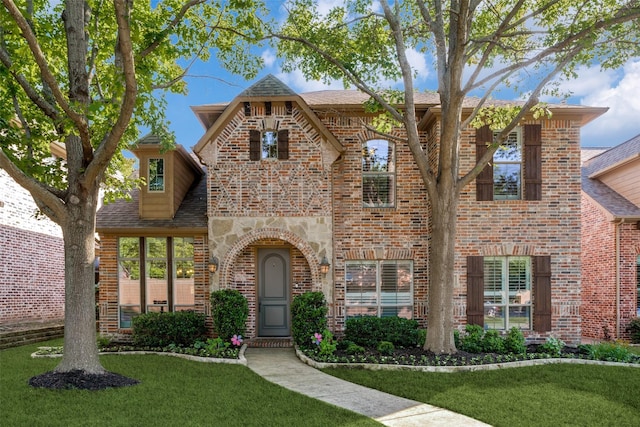 tudor home with brick siding, a front yard, a shingled roof, and stone siding