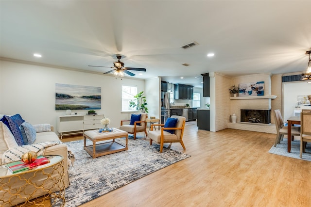 living area with ceiling fan, light wood-type flooring, a brick fireplace, ornamental molding, and recessed lighting