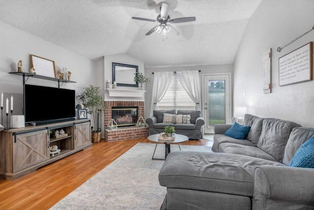 living room with lofted ceiling, light wood-style flooring, a textured ceiling, ceiling fan, and a brick fireplace