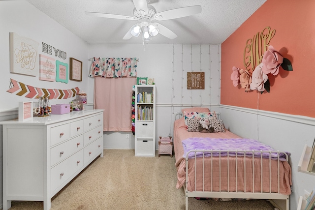 bedroom featuring a textured ceiling, light carpet, ceiling fan, and a wainscoted wall