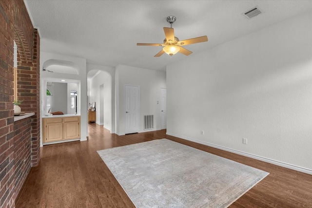 unfurnished living room featuring a ceiling fan, arched walkways, dark wood-style flooring, baseboards, and brick wall