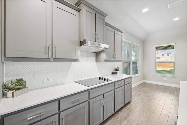 kitchen featuring gray cabinets, black electric cooktop, under cabinet range hood, baseboards, and backsplash