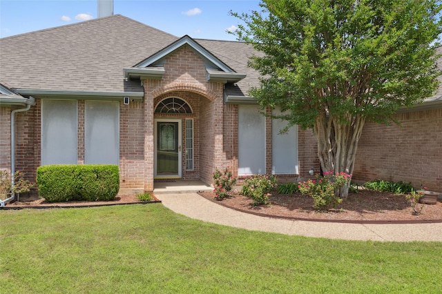 view of exterior entry featuring brick siding, roof with shingles, and a lawn